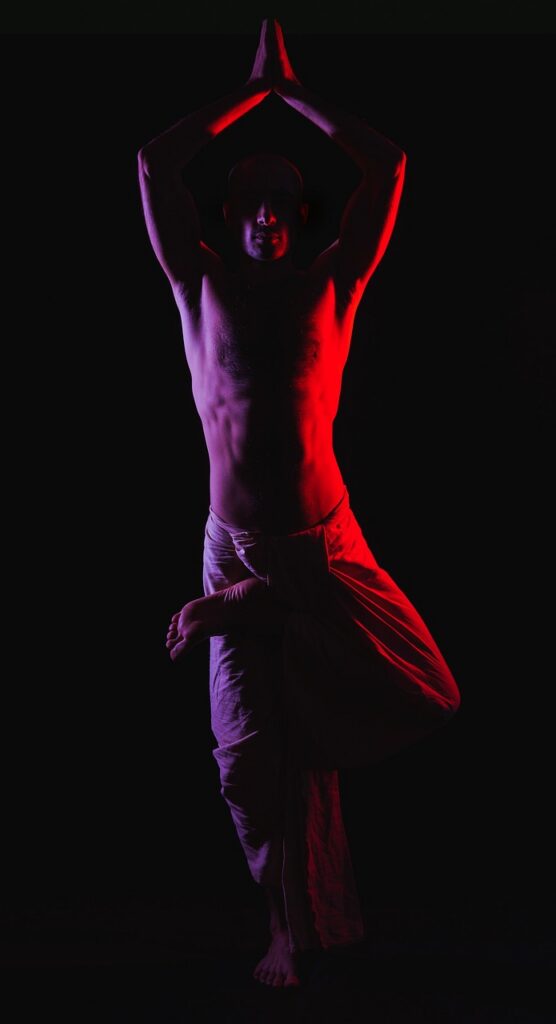 Miespuolinen paidaton joogi tekemässä joogausasentoa pimeässä mustalla taustalla. Häneen osuu oikealta punainen ja vasemmalta violetti valo. 