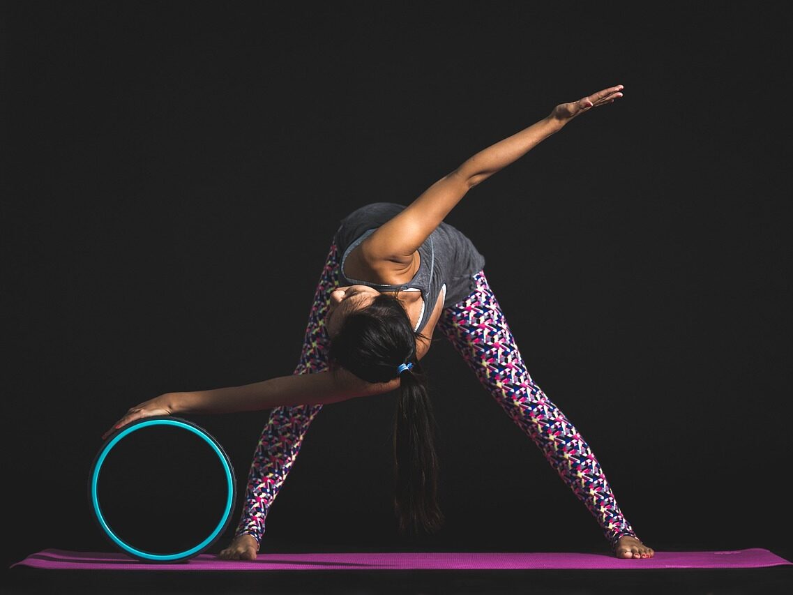 Naispuolinen joogaaja, joka venyttelee violetin värisellä joogamatolla. Hänen takanaan on musta tausta. Naisella on tummat pitkät hiukset, jotka ocat poninhännällä. Hänellä on harmaa toppi ja kirjavat urheilulegginssit.